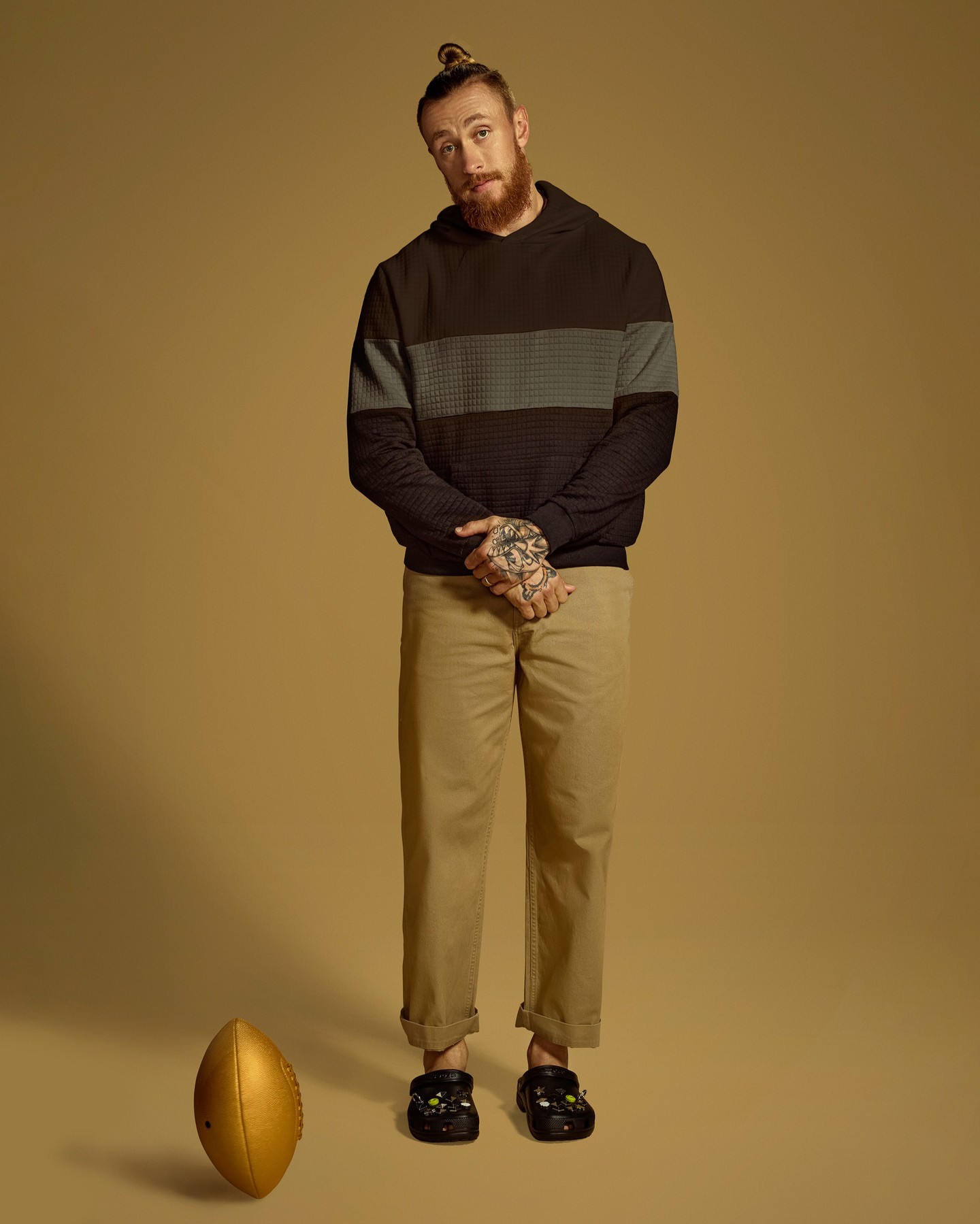Full studio shot of a man with a beard and top knot, wearing casual clothes and Crocs, standing next to a gold football on the floor.